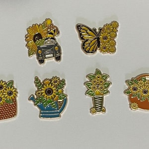 Może przedstawiać: Kolekcja emaliowanych przypinek z motywem słoneczników. Projekty obejmują słonecznik w koszu, motyla, konewkę, zabytkowy samochód i dekoracyjny wazon. Przypinki mają kolory żółty, zielony i brązowy z czarnymi obrysami.
