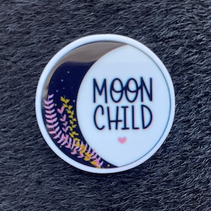 Könnte beinhalten: Ein runder, weißer Button mit dem Schriftzug "MOON CHILD" in schwarzer Schrift. Das Design umfasst einen Mond mit Sternenhimmel, rosa und gelbe Blumenakzente und ein kleines rosa Herz.