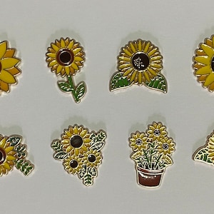 Könnte beinhalten: Eine Sammlung von acht Emaille-Ansteckern in Form von Sonnenblumen in verschiedenen Designs. Die Anstecker haben gelbe Blütenblätter, braune Zentren und grüne Blätter mit goldfarbenen Umrissen. Ein Anstecker zeigt Sonnenblumen in einem braunen Topf.