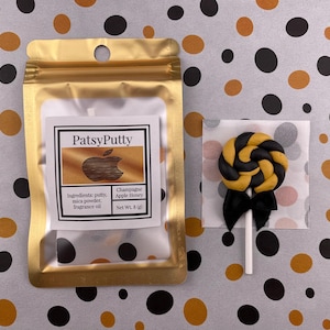 Könnte beinhalten: Ein goldener Metallic-Beutel mit einem Etikett "PatsyPutty" und einem schwarz-goldenen Lutscher auf einem weißen Stiel. Das Etikett listet Zutaten und Nettogewicht auf. Der Lutscher hat eine schwarze Schleife.
