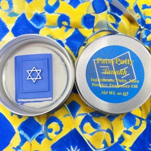 Könnte beinhalten: Zwei offene silberne Dosen auf einer blau-gelb gemusterten Oberfläche. Eine Dose enthält ein blaues Buch mit einem Davidstern. Die andere Dose hat ein blaues Etikett mit dem Text "Patsy Putty Tanakh" und Informationen zu den Inhaltsstoffen. Nettogewicht 10 (g).