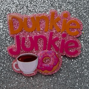 Könnte beinhalten: Ein pinkfarbener Glitzeraufkleber mit den Worten "Dunkie Junkie" in orangefarbenen und pinkfarbenen Buchstaben. Das Design umfasst eine weiße Kaffeetasse mit brauner Flüssigkeit und einen pinkfarbenen Donut mit Streuseln.