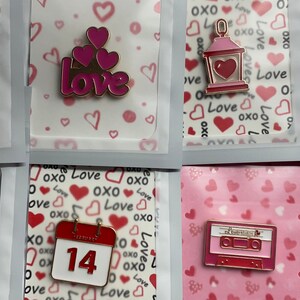 Könnte beinhalten: Eine Sammlung von Emaille-Anstecknadeln zum Valentinstag. Designs beinhalten das Wort "LOVE", Herzen, eine Laterne, einen Regenbogen, eine Geschenkbox, einen Kalender mit der Zahl 14 und eine Kassette. Jede Nadel befindet sich auf einer dekorativen Karte.