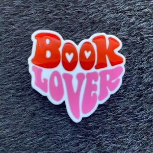 Może przedstawiać: Naklejka w kształcie serca z napisem "Book Lover" w stylu retro. Słowo "Book" jest w kolorze czerwonym z białymi obrysami, a "Lover" w kolorze różowym. Dwa małe białe serca znajdują się w słowie "Book".