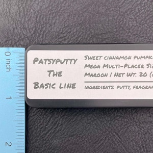Op de afbeelding: Een rechthoekige zwarte blikken doos met een wit etiket met de tekst Patsyputty The Basic Line, Sweet Cinnamon Pumpkin Mega Multi-Placer Size, Maroon / Net Wt. 20 (g) en Ingredients: Putty, Fragrance, naast een liniaal van 5,08 cm.