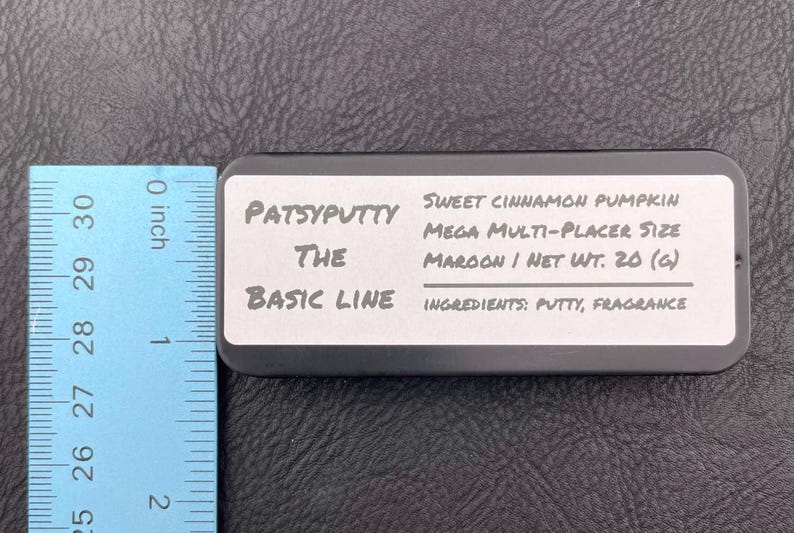 Op de afbeelding: Een rechthoekige zwarte blikken doos met een wit etiket met de tekst Patsyputty The Basic Line, Sweet Cinnamon Pumpkin Mega Multi-Placer Size, Maroon / Net Wt. 20 (g) en Ingredients: Putty, Fragrance, naast een liniaal van 5,08 cm.