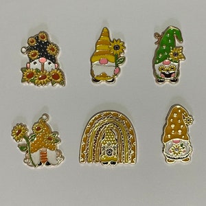 Może przedstawiać: Kolekcja sześciu kolorowych zawieszek z krasnalami z motywami słoneczników i tęczy. Zawieszki mają różne kolory, w tym żółty, zielony i złoty, ze skomplikowanymi detalami i metalicznym wykończeniem. Idealne do tworzenia biżuterii.