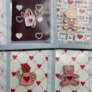 Könnte beinhalten: Eine Sammlung von Emaille-Anstecknadeln zum Valentinstag. Designs umfassen einen Ring in einer herzförmigen Schachtel, eine Gießkanne mit einem Herz, einen Teddybären mit "I Love You" und einen Briefkasten. Weitere Anstecknadeln zeigen einen Blumenstrauß, einen Teddybären und einen Umschlag mit Herzen.