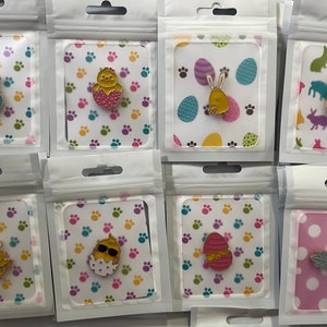 Könnte beinhalten: Eine Sammlung von Emaille-Anstecknadeln zum Thema Ostern, jeweils verpackt in einer transparenten Plastiktüte. Die Anstecknadeln zeigen Designs wie Küken, Hasen und Ostereier. Die Hintergrundkarten haben Pfotenabdrücke, Eier und Hasenmuster.