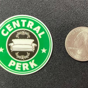 Pu&ograve; includere: Un adesivo rotondo verde con le parole "CENTRAL PERK" in bianco, che circondano un'immagine in bianco e nero di un divano. Una moneta da un quarto di dollaro americano &egrave; accanto per la scala.