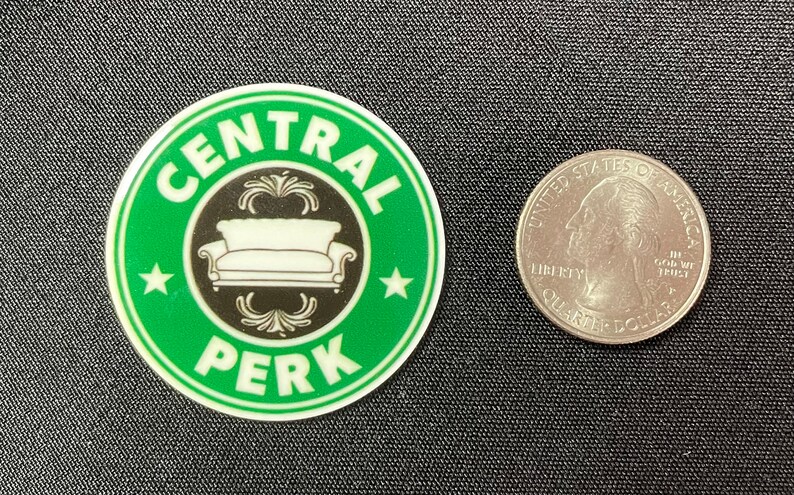 Pu&ograve; includere: Un adesivo rotondo verde con le parole "CENTRAL PERK" in bianco, che circondano un'immagine in bianco e nero di un divano. Una moneta da un quarto di dollaro americano &egrave; accanto per la scala.