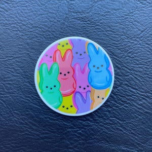 Könnte beinhalten: Ein runder Aufkleber mit bunten Peeps-Marshmallow-Hasen in Grün, Rosa, Lila, Blau und Gelb. Die Hasen haben kleine schwarze Augen und sind auf einem rosa Hintergrund angeordnet.