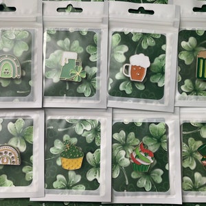 Könnte beinhalten: Eine Sammlung von Emaille-Anstecknadeln zum St. Patrick's Day in transparenten Plastiktüten. Die Pins zeigen einen Regenbogen, Bierkrüge, einen Cupcake und einen Kaktus, alle in Grün und Gold. Der Hintergrund ist ein grünes Kleeblattmuster.