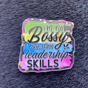 Könnte beinhalten: Ein farbenfroher rechteckiger Anstecker mit dem Text "I'm not Bossy I just have leadership SKILLS". Der Anstecker hat ein lebendiges Design mit grünen, gelben, blauen und rosa Hintergründen im Aquarellstil. Der Anstecker ist mit einem schwarzen Rand und Glitzer versehen.