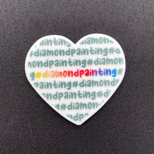 Könnte beinhalten: Ein weißer, herzförmiger Anhänger mit dem Text "#diamondpainting" in verschiedenen Farben und Schriftarten. Der Anhänger ist vor einem dunkelgrauen Hintergrund platziert, der den Text hervorhebt.