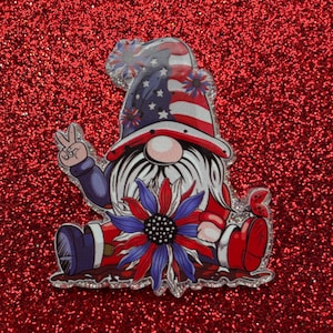 Può includere: Illustrazione di uno gnomo patriottico con un segno di pace, che indossa un cappello e tiene un girasole. Il cappello presenta il disegno della bandiera americana con stelle e strisce. L'abito dello gnomo è rosso, bianco e blu.