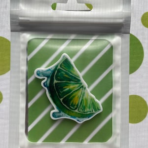 Könnte beinhalten: Ein Limettenscheiben-Aufkleber mit Aquarelldetails, verpackt in einer transparenten Plastiktüte. Der Aufkleber zeigt einen grünen Limettenschnitz mit blauen Akzenten, vor einem grün-weiß gestreiften Hintergrund.