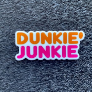 Könnte beinhalten: Ein weißer, rechteckiger Anhänger mit den Worten "DUNKIE' JUNKIE" in orangefarbenen und pinkfarbenen Blockbuchstaben. Der Anhänger befindet sich vor einem strukturierten, grauen Hintergrund.