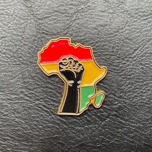 Peut inclure: Une épingle dorée représentant le continent africain, avec un poing noir et les couleurs rouge, jaune et vert. L'épingle est posée sur un fond noir texturé.