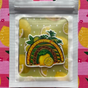 Könnte beinhalten: Eine transparente Plastiktüte mit einem Aufkleber mit Regenbogenmuster. Der Regenbogen hat gelbe, grüne und braune Streifen mit einem Leopardenmuster. Der Aufkleber zeigt auch Zitronen und grüne Blätter. Der Hintergrund in der Tüte hat ein Zitronenmuster.