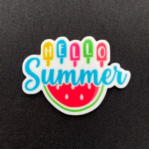 Puede incluir: Un charm blanco con temática de verano con un diseño de rodaja de sandía y helado. El texto "HELLO SUMMER" está escrito en azul y letras coloridas. El charm probablemente está hecho de plástico o resina.