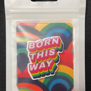 Puede incluir: Una pegatina cuadrada con el texto "BORN THIS WAY" en un diseño colorido y en capas. El fondo presenta un patrón de arcos iris. La pegatina está empaquetada en una bolsa de plástico transparente con un agujero para colgar.