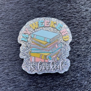 Könnte beinhalten: Ein glitzernder Anstecker mit den Worten "My Weekend is Booked" in Pastellfarben. Der Anstecker zeigt einen Stapel Bücher in Blau, Gelb und Rosa mit einem blauen Lesezeichen.