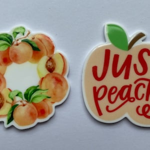 Könnte beinhalten: Zwei Aufkleber zum Thema Pfirsich. Einer ist ein Kranz aus Pfirsichen mit grünen Blättern. Der andere ist ein apfelförmiger Aufkleber mit den Worten "JUST peachy" in roter Schrift. Die Aufkleber haben einen weißen Hintergrund.