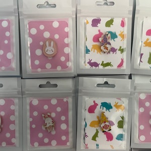Könnte beinhalten: Oster-Emaille-Anstecknadeln in transparenter Kunststoffverpackung. Designs umfassen Ostereier, Hasen und Zwergcharaktere. Die Anstecknadeln werden auf rosa Karten mit weißen Punkten und Karten mit Hasenmuster präsentiert.