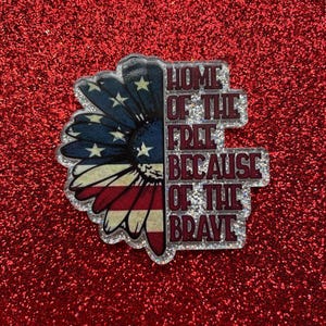 Puede incluir: Pin patriótico con un diseño de girasol, con los colores de la bandera estadounidense: rojo, blanco y azul. El texto "HOME OF THE FREE BECAUSE OF THE BRAVE" se muestra en una combinación de colores a juego.