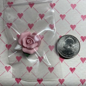Könnte beinhalten: Eine rosa Rose aus Ton, mit weißen und grünen Akzenten, wird in einer transparenten Plastiktüte präsentiert. Eine Münze von 25 Cent ist neben der Rose zum Größenvergleich. Der Hintergrund ist ein Muster aus rosa Herzen.