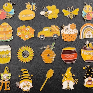Könnte beinhalten: Eine Sammlung bunter Emaille-Anstecknadeln mit einem Bienen- und Honig-Thema. Designs umfassen Bienen, Waben, Honigtöpfe, eine Sonnenblume, einen Lastwagen, einen Regenbogen und Zwerge. Die Pins sind in Gelb-, Orange-, Schwarz- und Weißtönen gehalten.