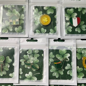 Könnte beinhalten: Eine Sammlung von Emaille-Anstecknadeln zum St. Patrick's Day in transparenten Plastiktüten. Die Anstecknadeln zeigen ein Haus, eine Goldmünze mit einem Kleeblatt, ein Herz mit der italienischen Flagge, einen Lolli, grüne Sonnenbrillen, eine Pfeife und ein Hufeisen mit "Good Luck".