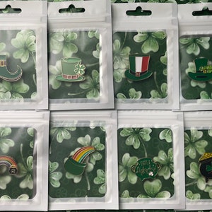 Könnte beinhalten: Eine Sammlung von St. Patrick's Day Emaille-Anstecknadeln in transparenten Kunststoffverpackungen. Die Anstecknadeln zeigen verschiedene Designs, darunter einen grünen Stiefel, Zylinder, einen Regenbogen, einen Goldtopf und ein Kleeblatt. Der Hintergrund ist ein grünes Kleeblattmuster.
