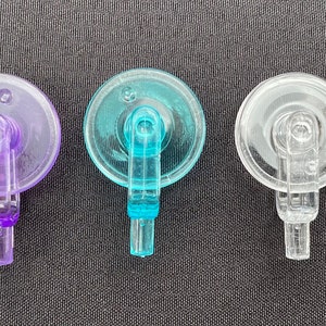 Könnte beinhalten: Fünf transparente Kunststoff-Saugnäpfe in einer Reihe, in Gelb, Lila, Türkis, Klar und Rosa. Jede hat einen kreisförmigen Deckel und einen kleinen Griff. Sie liegen auf einer dunkelgrauen Oberfläche.