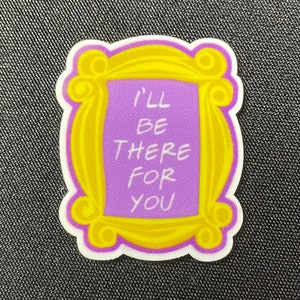 Pu&ograve; includere: Un adesivo a tema Friends con una cornice gialla e un centro viola. Il testo "I'll be there for you" &egrave; scritto in bianco. L'adesivo &egrave; su una superficie strutturata grigio scuro.