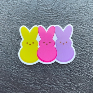 Könnte beinhalten: Eine farbenfrohe Osterdekoration mit drei Marshmallow-Hasenfiguren. Die Hasen sind gelb, rosa und lila, jeder mit kleinen braunen Punktaugen. Die Figuren haben einen weißen Rand und sind vor einem dunklen Hintergrund platziert.