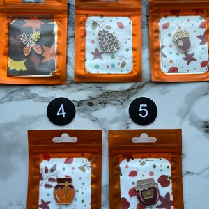 Könnte beinhalten: Fünf Emaille-Anstecknadeln in orangefarbener Verpackung. Die Anstecknadeln zeigen herbstliche Motive: Eichenblätter und Eicheln, einen Tannenzapfen, eine Eichel und Gläser mit Honig und Marmelade. Der Hintergrund jeder Anstecknadel ist ein herbstliches Muster mit Blättern.