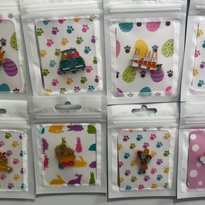 Könnte beinhalten: Eine Sammlung von Emaille-Anstecknadeln zum Thema Ostern in transparenter Kunststoffverpackung. Die Anstecknadeln zeigen Designs wie einen LKW mit Ostereiern, Hasen und einem Regenbogen. Die Rückkarten haben bunte Muster von Eiern, Pfotenabdrücken und Hasen.