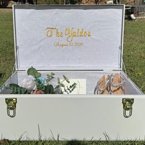 May include: A white wooden keepsake box with gold-coloured hardware, open to reveal a floral arrangement, a card, and a pair of embellished sandals. The box's interior is lined with white fabric, and the lid features gold embroidered text: "The Yaldos August 22, 2026."