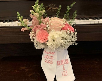 May include: A floral arrangement featuring pink roses, carnations, and white hydrangeas with green stems. Two white ribbons hang from the flowers, with pink text reading Charter HOCO 2025 on one and BRAYLEN & LILY on the other, set against a dark piano.