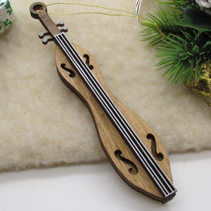 May include: Wooden dulcimer Christmas ornament with black and white strings. The ornament is brown with a light-colored center. A small wrapped gift, pinecone, and holiday greenery are in the background.