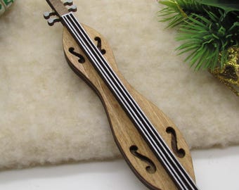 May include: Wooden dulcimer Christmas ornament with black and white strings. The ornament is brown with a light-colored center. A small wrapped gift, pinecone, and holiday greenery are in the background.