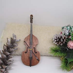 May include: A wooden double bass ornament with a brown stain finish and a gold string. The ornament is hanging from a white string.