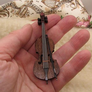 May include: A miniature wooden cello with a bow, featuring a dark brown body and black details. The cello has white strings and a red accent on the scroll. The instrument is a small decorative item.