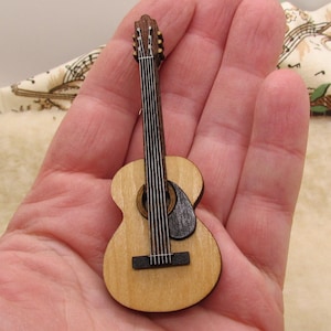May include: A miniature acoustic guitar made of light-colored wood, featuring a dark sound hole and fretboard. The guitar has six strings and a detailed headstock. The instrument is held in a hand, showcasing its small size.