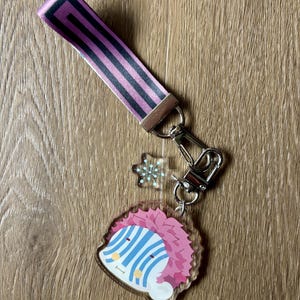 May include: A keychain set featuring a pink and black striped wrist strap, a silver-colored clasp, a snowflake charm, and a clear acrylic charm with a cartoon character design. The character has pink hair, blue stripes, and a smiling face.