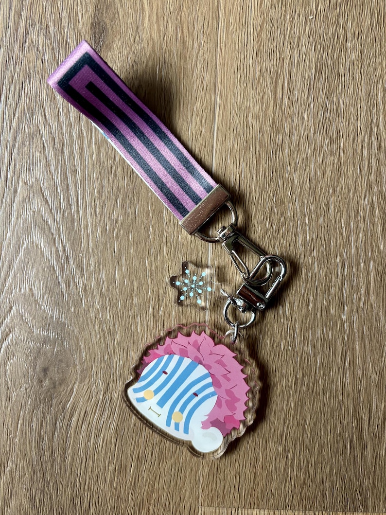 May include: A keychain set featuring a pink and black striped wrist strap, a silver-colored clasp, a snowflake charm, and a clear acrylic charm with a cartoon character design. The character has pink hair, blue stripes, and a smiling face.