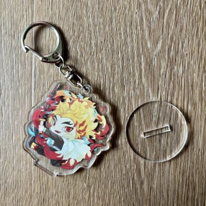 May include: A clear acrylic keychain featuring a colorful anime character with yellow hair and a red and black outfit. The keychain has a silver metal clasp and a small clear stand.