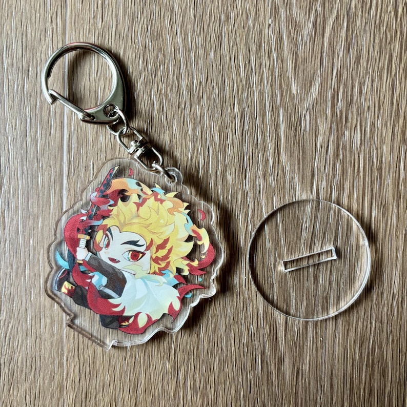 May include: A clear acrylic keychain featuring a colorful anime character with yellow hair and a red and black outfit. The keychain has a silver metal clasp and a small clear stand.